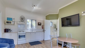 A compact kitchen with white appliances, a dining area with a table and chairs, and a flat-screen TV mounted on the wall. - Port Huon Cottages (Port Huon)