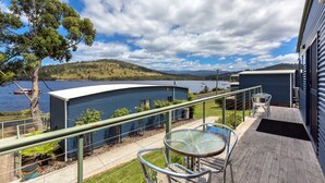 A waterfront property with a blue building, a balcony with a glass table and chairs, and a view of the lake and hills. - Port Huon Cottages (Port Huon)