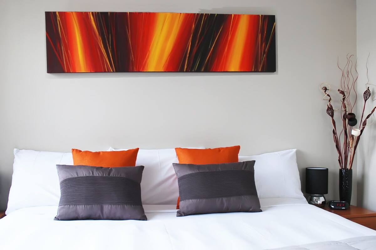 Habitación de hotel con una cama grande, dos almohadas, un cuadro abstracto en la pared, un jarrón con plantas secas y una mesita de noche con una lámpara y un reloj.