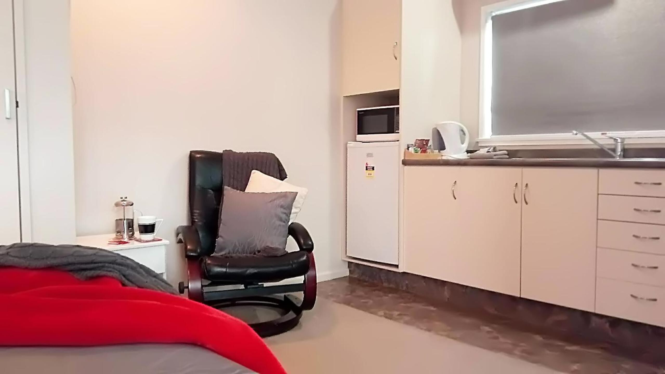 A compact room with a bed, a black chair with cushions, a small table with a cup and a kettle, a microwave, and a kitchenette with cabinets.