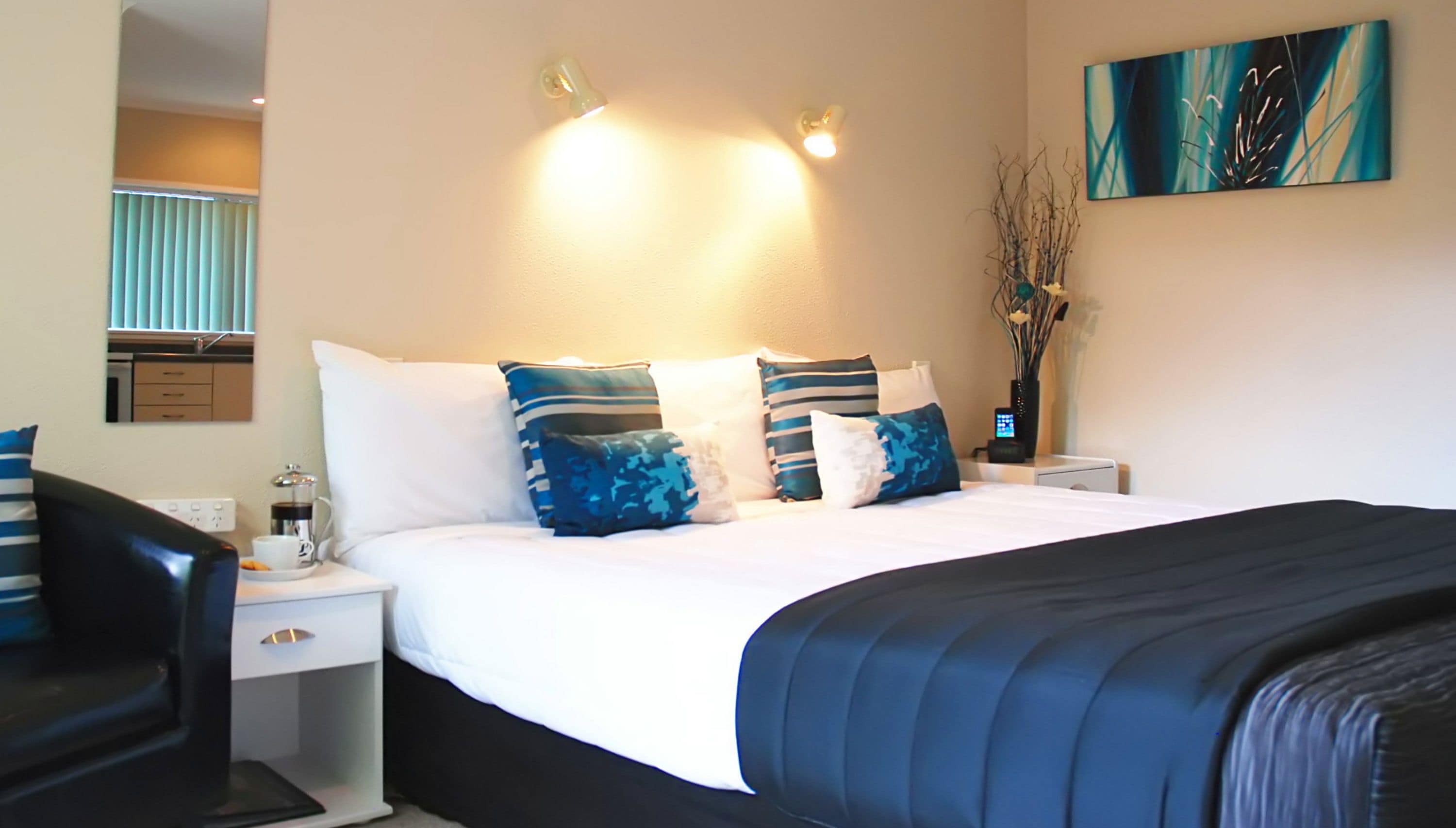 A hotel room with a bed, bedside tables, a nightstand, a lamp, a vase with branches, and a painting on the wall.