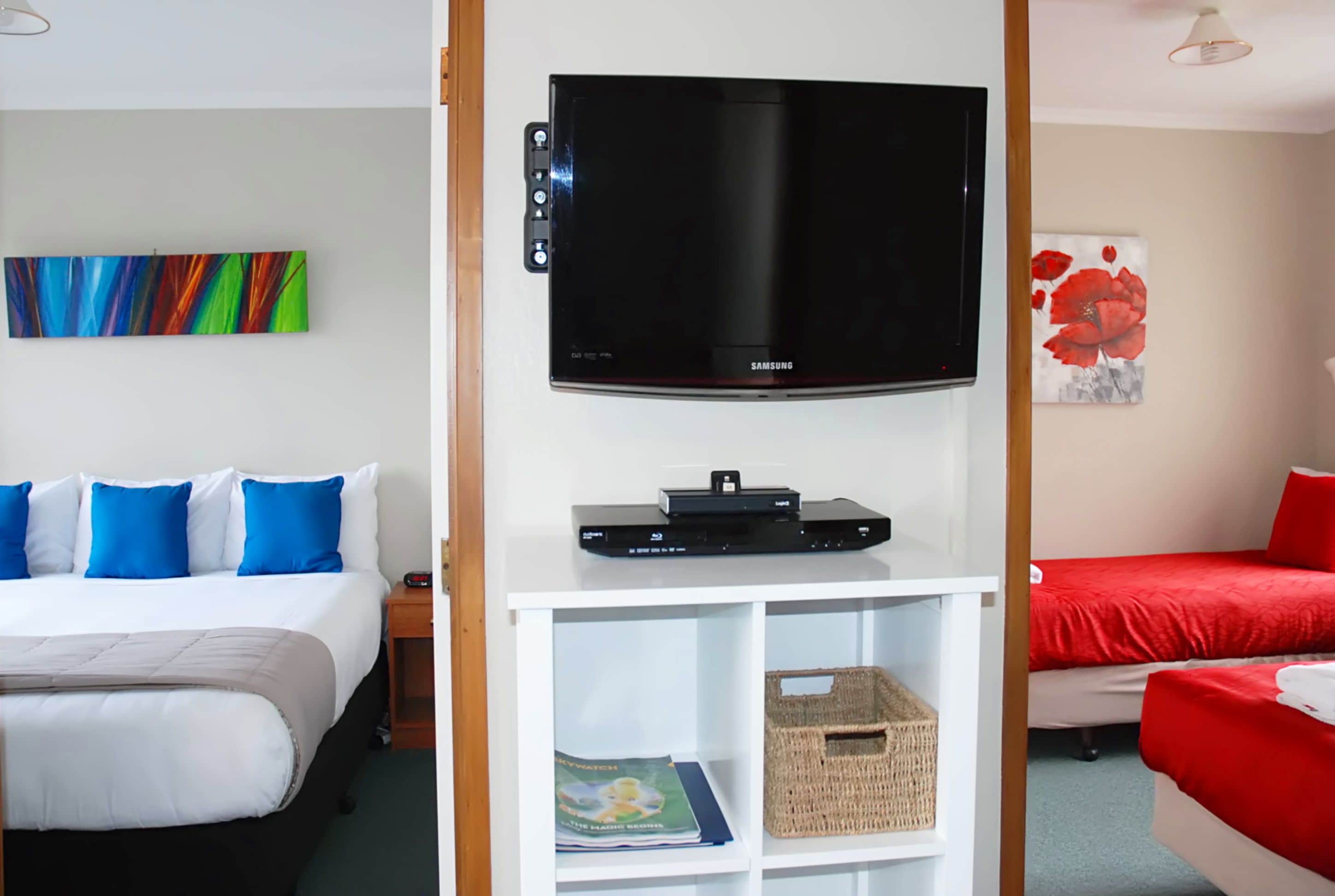 A hotel room with a bed, a TV on a stand, a painting on the wall, and a red bedspread.