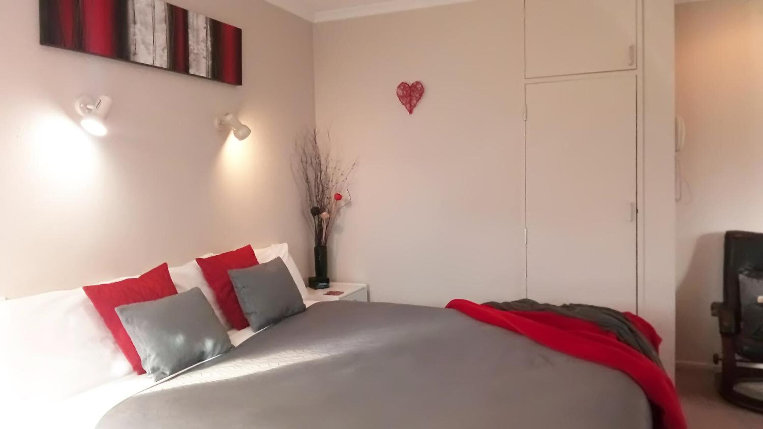 A bedroom with a bed, bedside tables, a nightstand, a vase with branches, and a heart-shaped decoration on the wall.