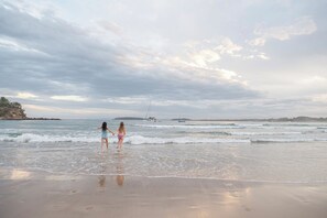 Eine Strandansicht mit zwei Personen, die im flachen Wasser stehen, Booten in der Ferne und einer felsigen Küste.