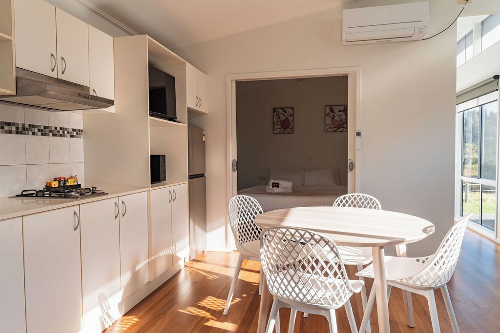 A modern kitchen with white cabinetry, a dining area with a white table and chairs, and a view through an open door to a bedroom.