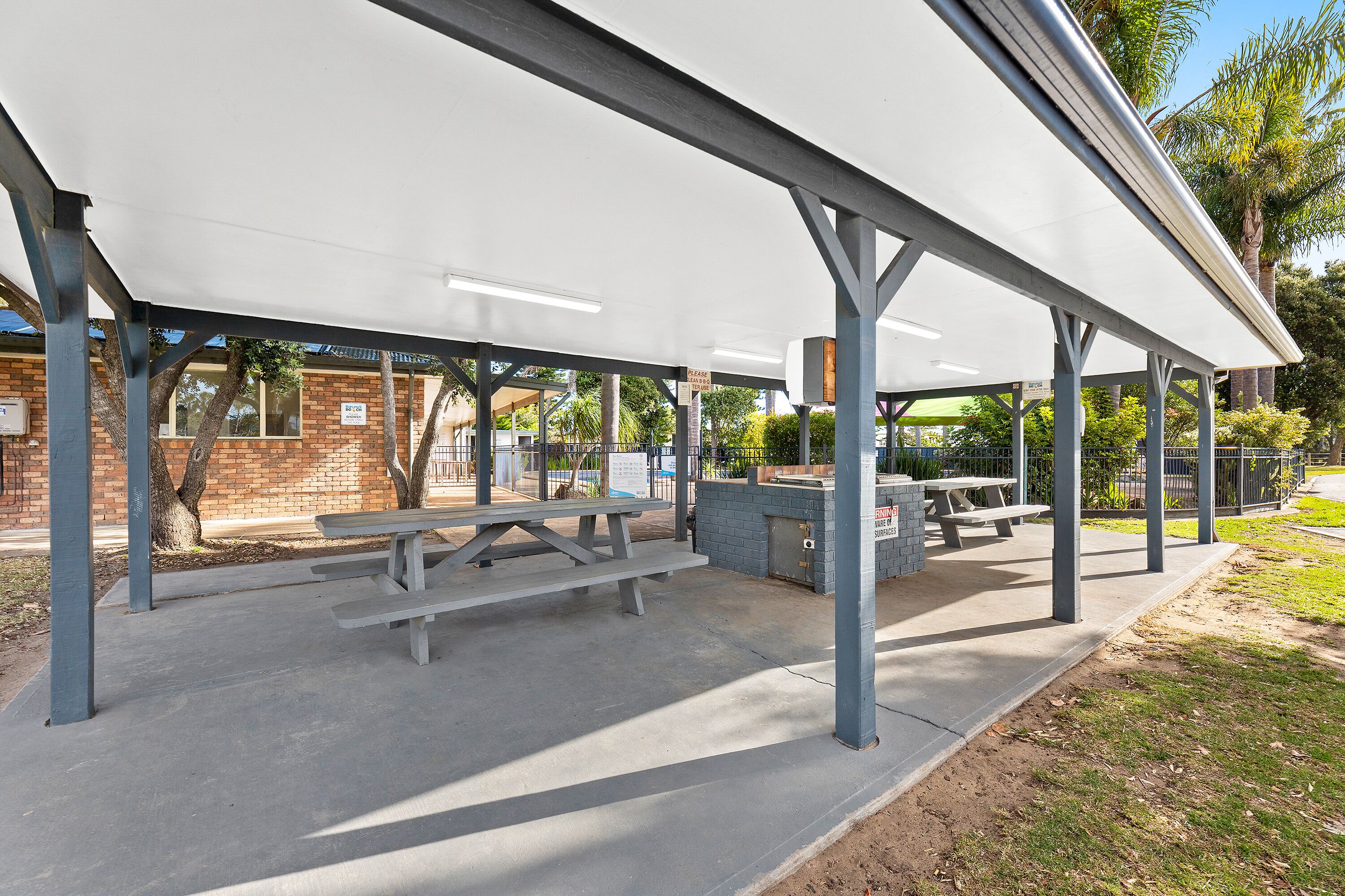 A covered picnic area with benches and a table, surrounded by a brick building and palm trees.