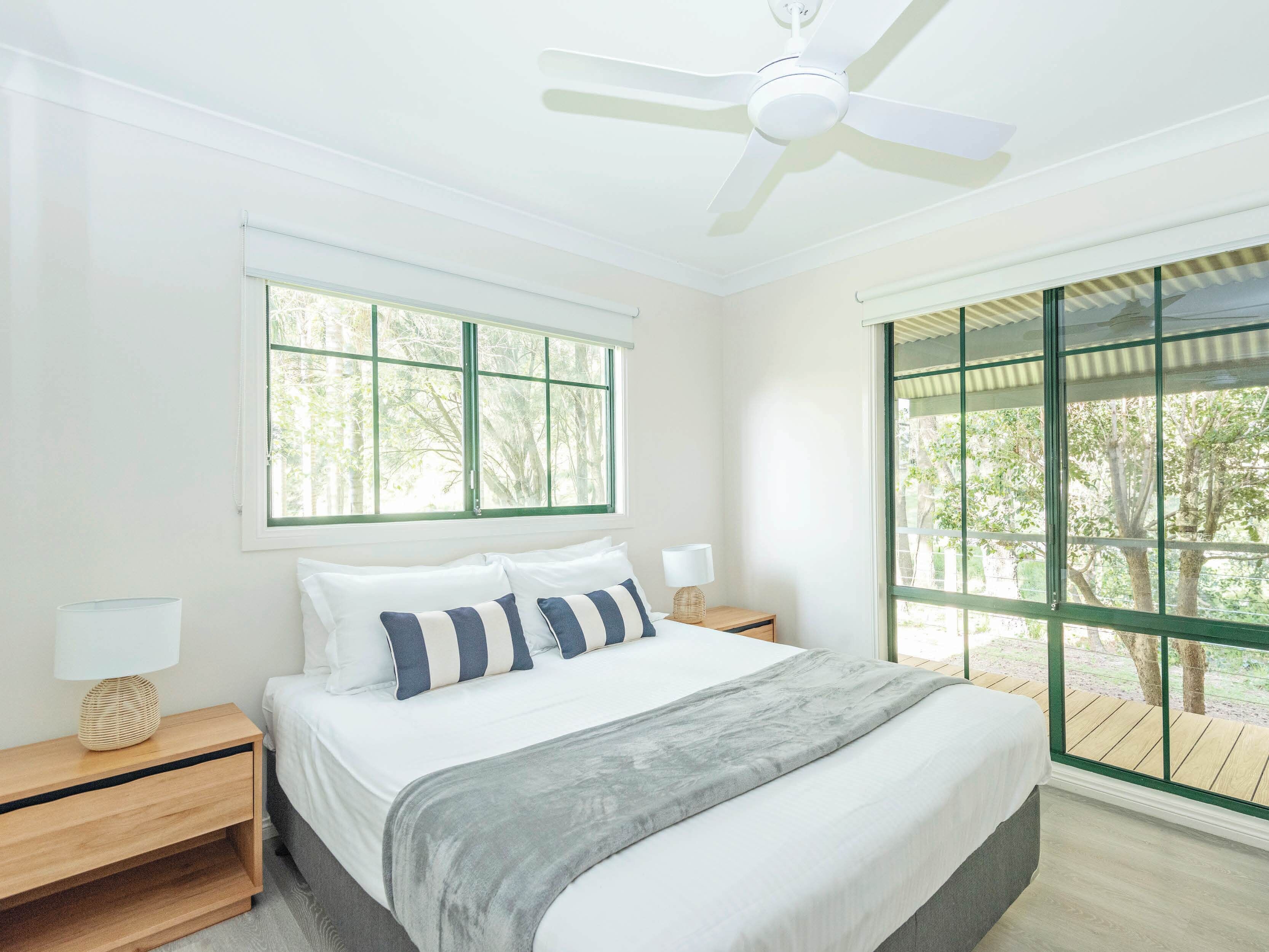 Ein Schlafzimmer mit Bett, Nachttischen, Deckenventilator und großen Fenstern mit Blick auf Bäume.