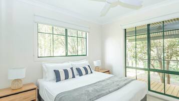 Ein Schlafzimmer mit Bett, Nachttischen, Deckenventilator und großen Fenstern mit Blick auf Bäume.