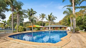 Una piscina con solárium de piedra, rodeada de palmeras y una casa al fondo.