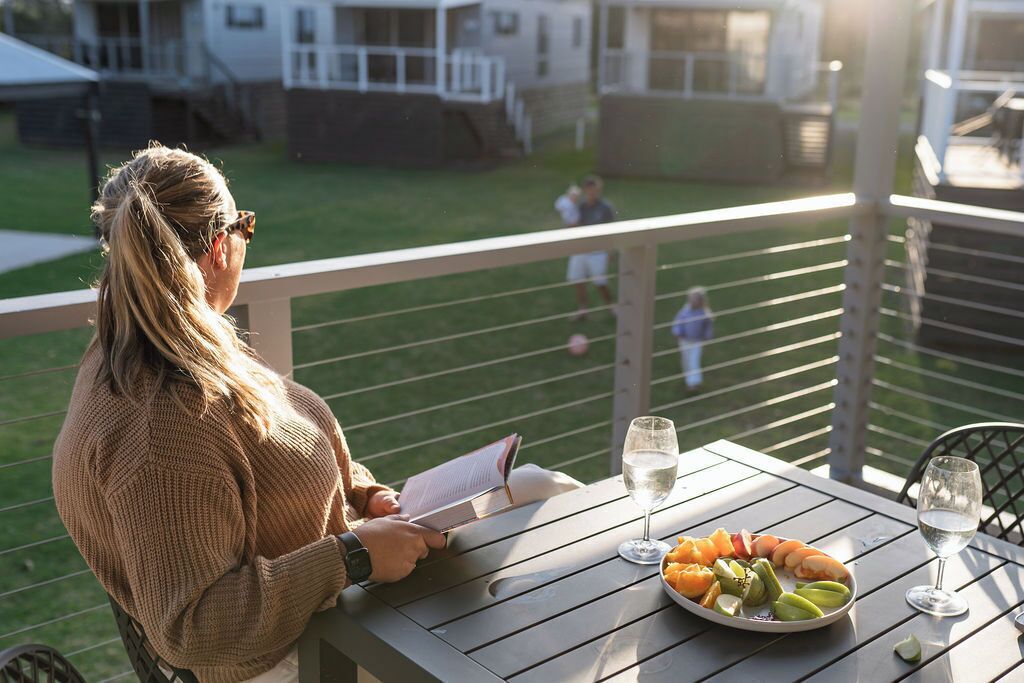Eine Person liest ein Buch auf einem Balkon, an dem ein Tisch mit Meeresfrüchten und Weingläsern gedeckt ist.