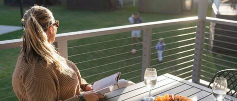 Eine Person liest ein Buch auf einem Balkon, an dem ein Tisch mit Meeresfrüchten und Weingläsern gedeckt ist.