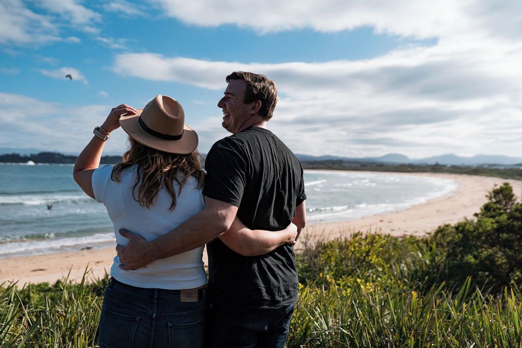 A couple standing by a beach with a view of the ocean and distant mountains.