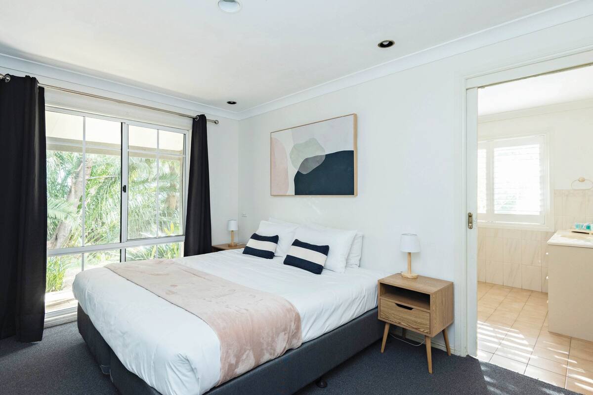 Une chambre à coucher avec un lit, une table de chevet, une fenêtre avec des rideaux, un tableau au mur et une salle de bain visible à travers une porte ouverte.