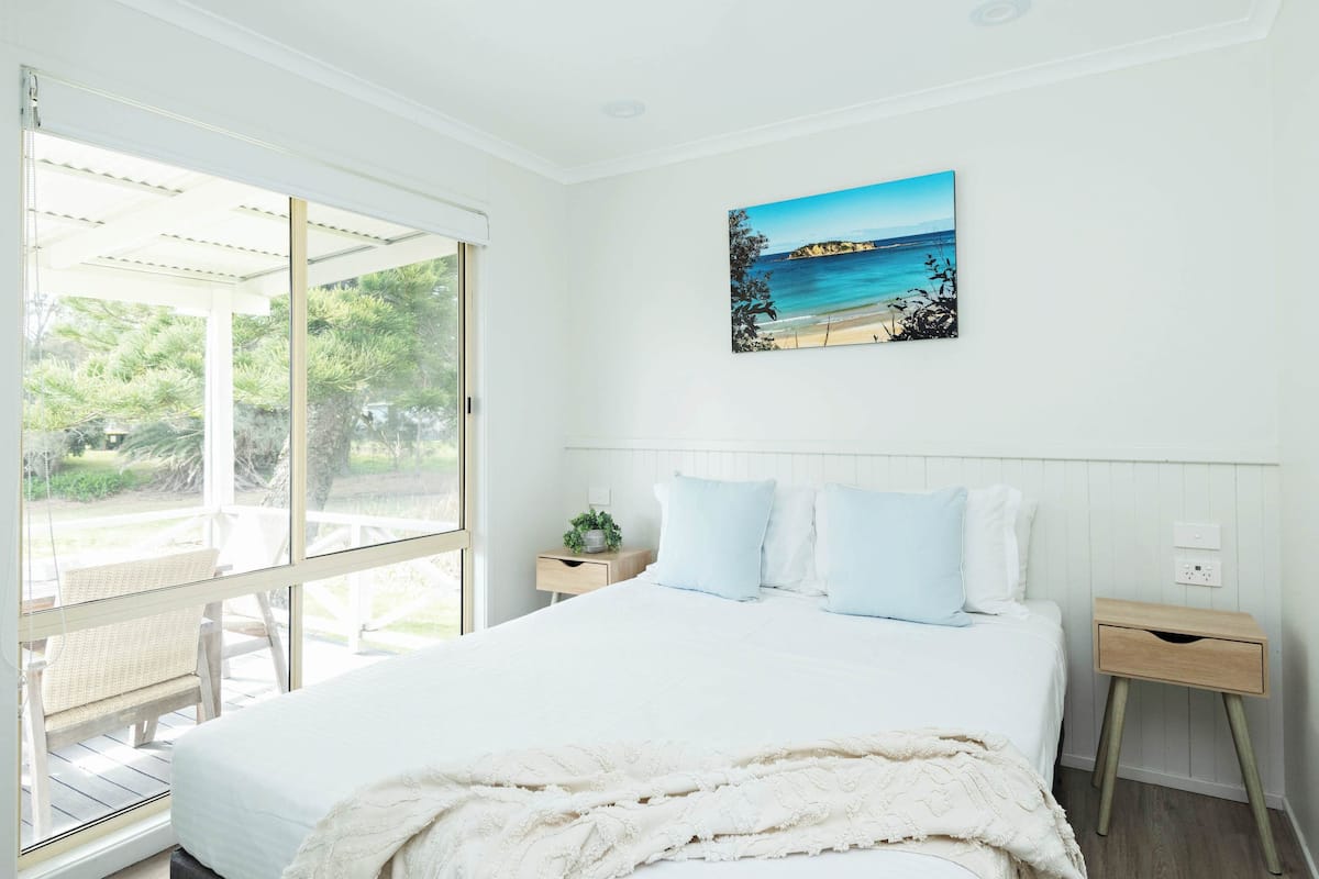 Une chambre avec un grand lit, une fenêtre donnant sur un paysage verdoyant, une table de chevet et un tableau accroché au mur.