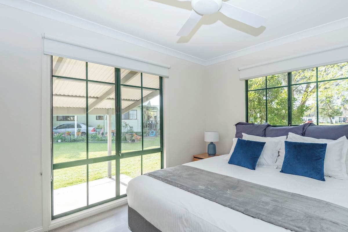 Une chambre avec un grand lit, un ventilateur de plafond, une table de chevet et une fenêtre donnant sur un jardin et une maison.