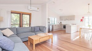 Una sala de estar moderna con un sofá seccional gris, una mesa de centro de madera y una cocina con armarios blancos y una mesa de comedor.