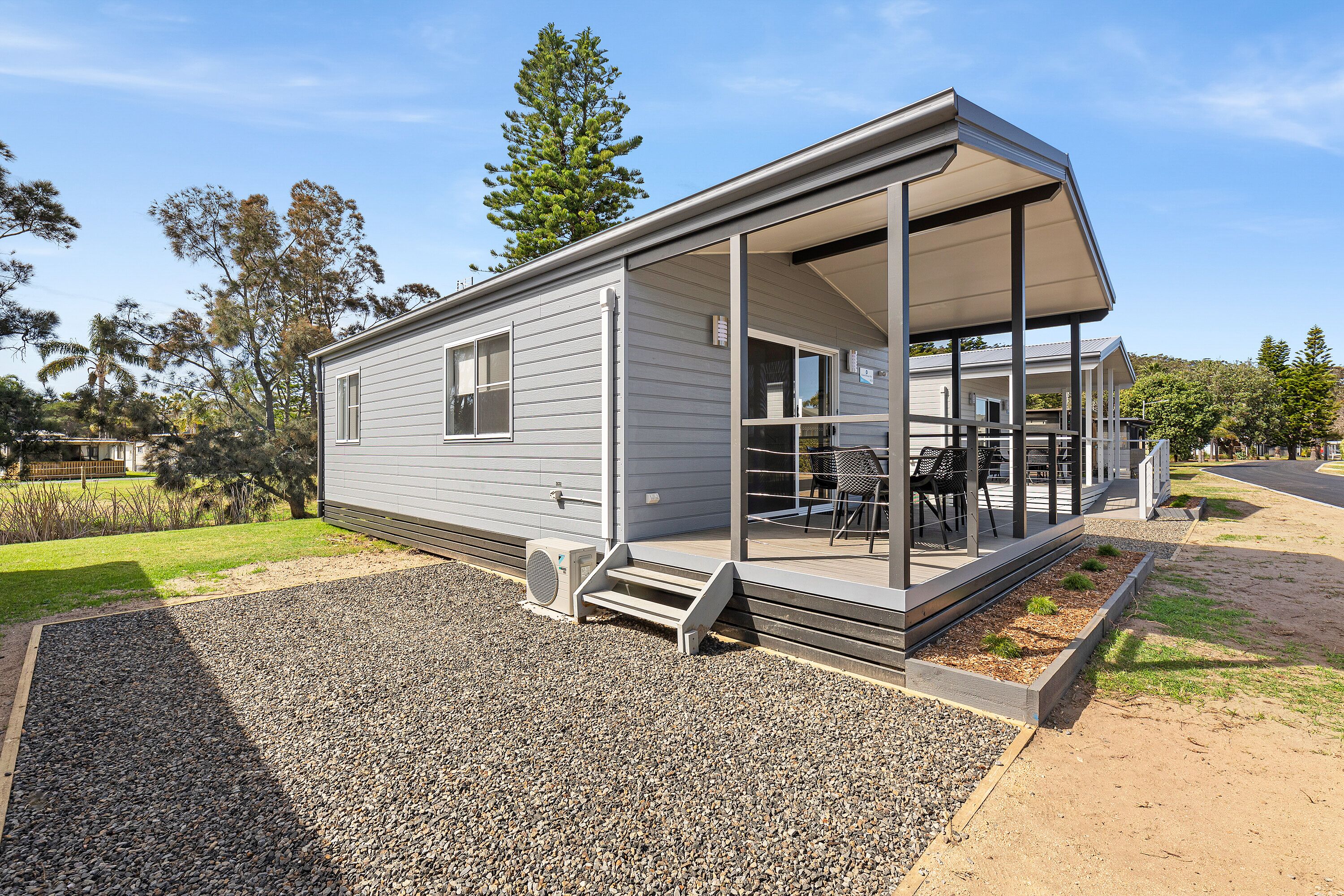 A modern single-story mobile home with a covered porch, outdoor seating, and a gravel driveway.