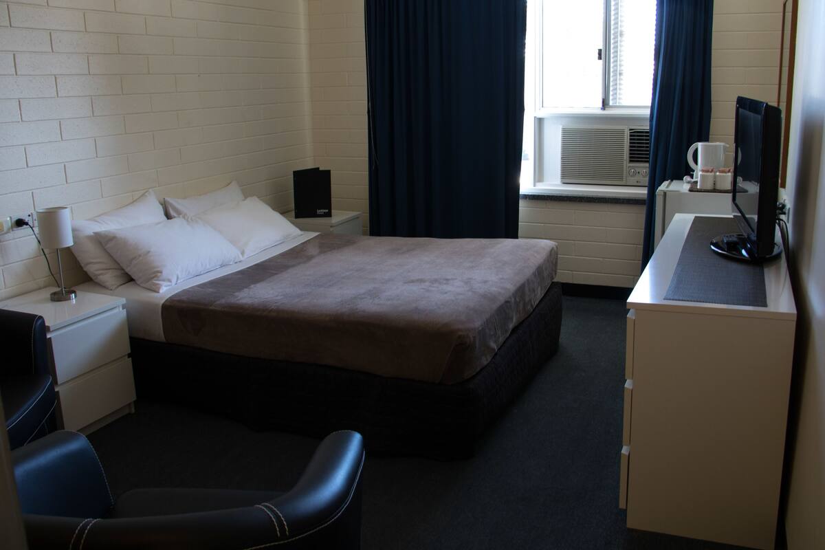 A hotel room with a bed, a desk with a computer, a chair, a nightstand, a lamp, and a window with curtains.