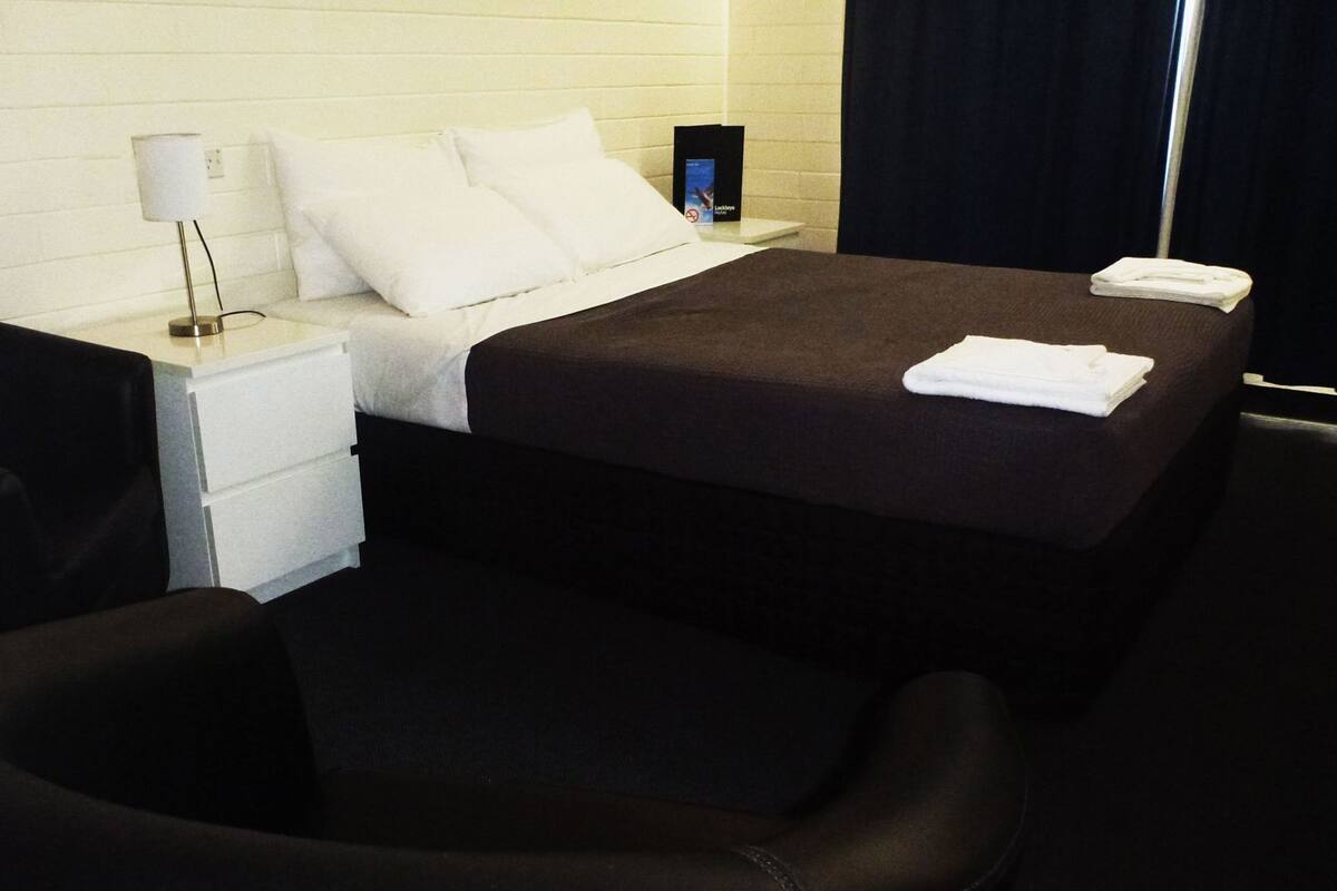 A hotel room with a bed, a nightstand, a lamp, a chair, and a folded towel.
