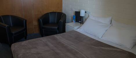 Habitación de hotel con una cama, dos sillones de cuero negro, una mesita de noche con una lámpara y una pared de ladrillo blanco.