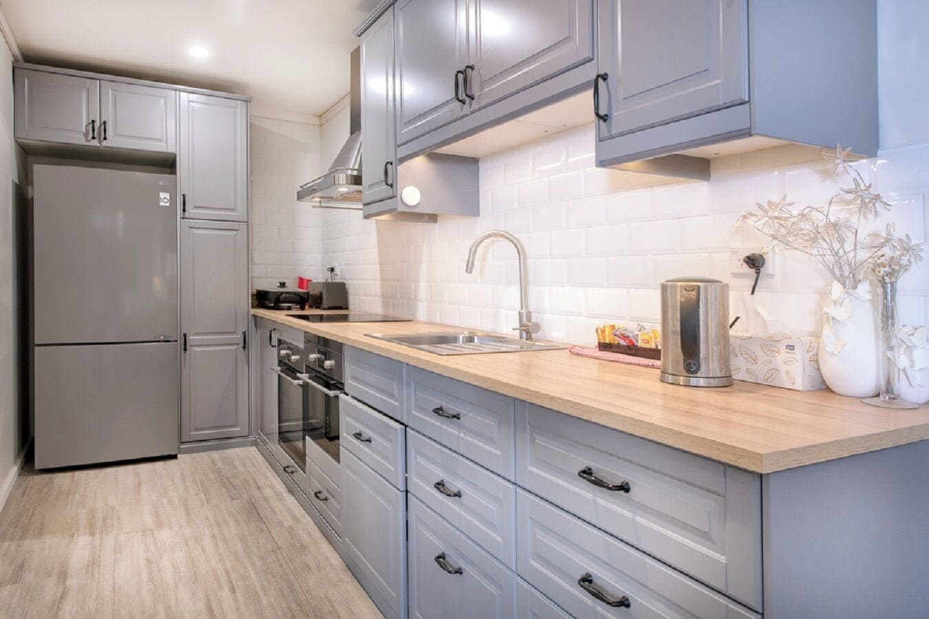 Une cuisine moderne avec des meubles gris, un réfrigérateur en acier inoxydable, un four encastré et un plan de travail en bois.