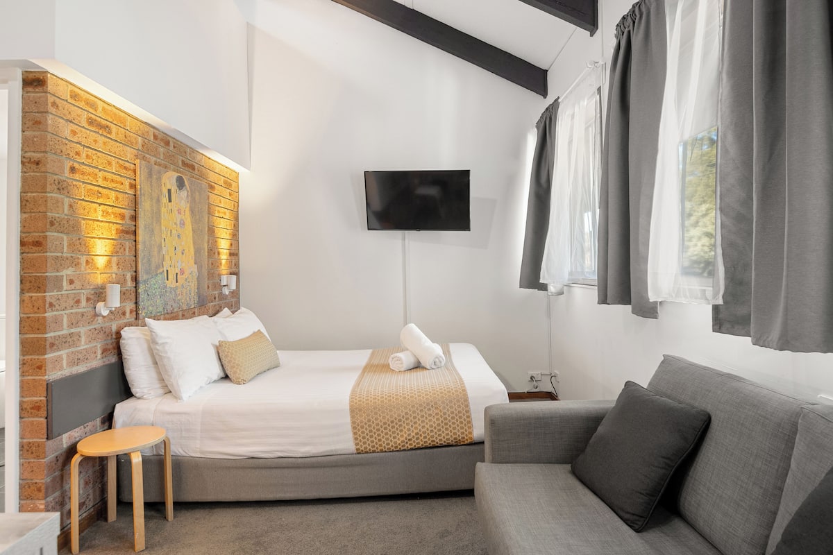 A modern hotel room with a brick accent wall, a flat-screen TV mounted on the wall, a bed with a white and beige bedspread, a grey sofa, and a small wooden stool.
