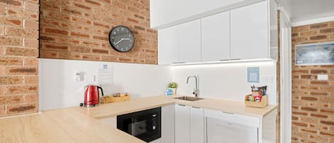 Una cocina moderna con pared de ladrillo, armarios blancos, lavavajillas negro, hervidor rojo y un reloj en la pared.