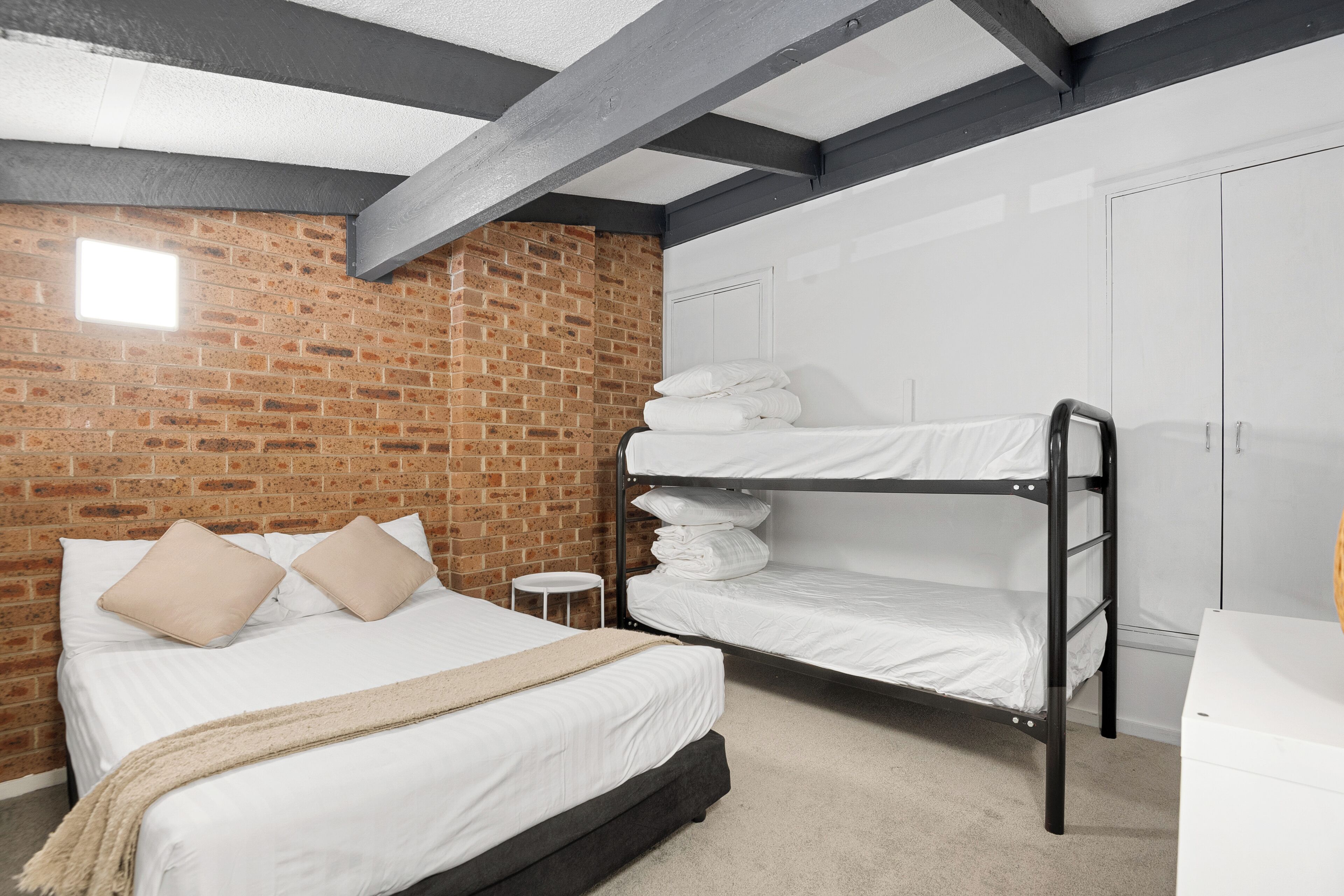 A bedroom with a brick wall, a bunk bed, a single bed with pillows, and a white wardrobe.