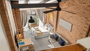 A loft-style bedroom with a brick wall, a ceiling fan, a bed with white linens, a wooden table, and a view of the outside.