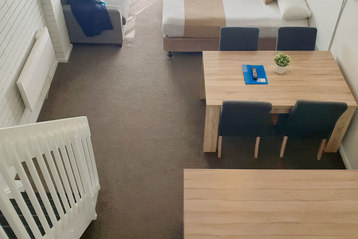 A hotel room with a bed, a seating area with a table and chairs, a small table with a plant, a white radiator, and a wall-mounted TV.