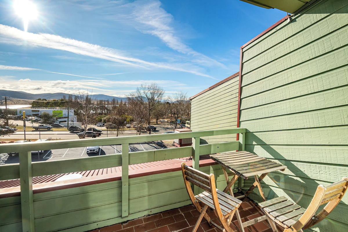 A balcony with a wooden table and chairs, overlooking a parking lot and distant mountains.