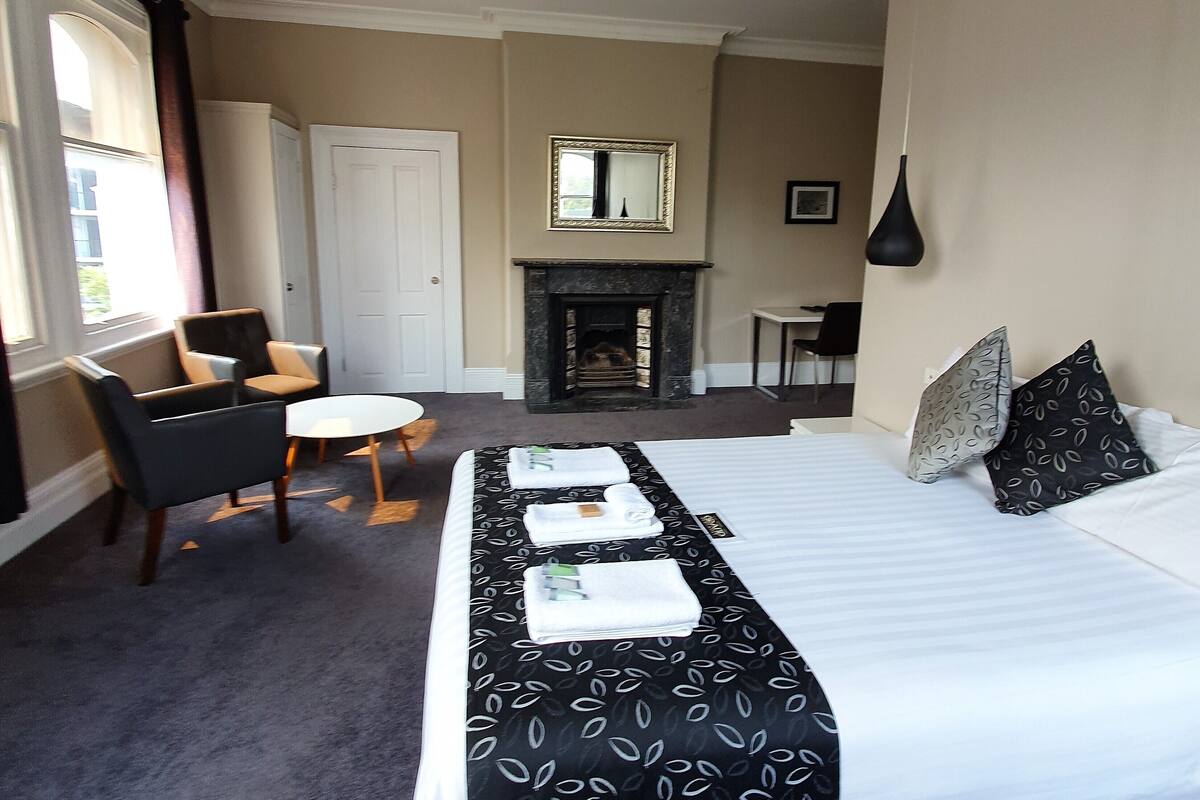Habitación de hotel con una cama grande, una mesita redonda pequeña, dos sillas, una chimenea y un escritorio con una silla.