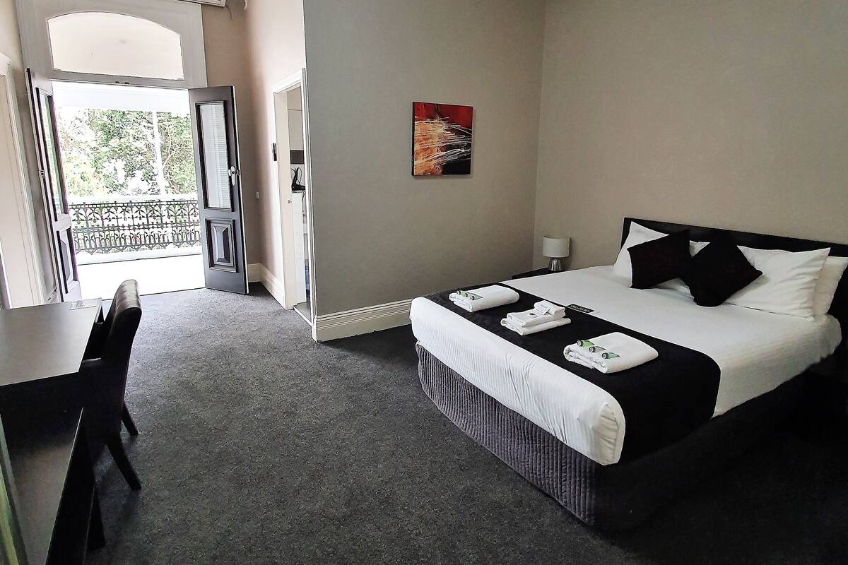Habitación de hotel con cama, escritorio y silla. Tiene un balcón con barandilla y una puerta que da al exterior.