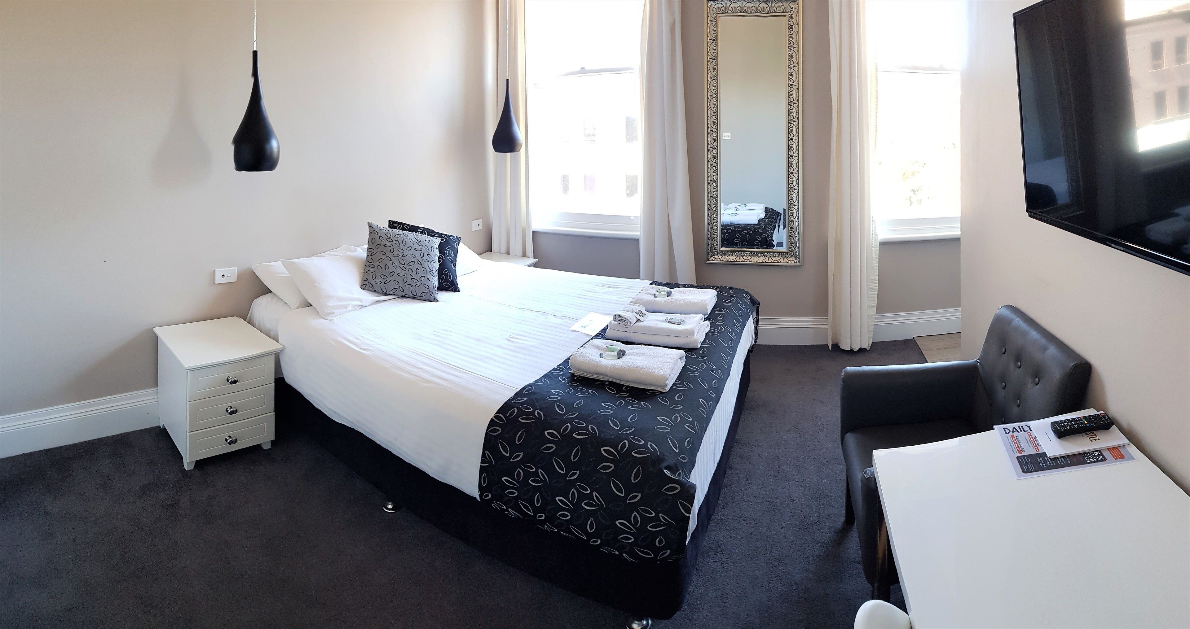 A hotel room with a neatly made bed, a bedside table with a remote control, a chair, and a large mirror on the wall.