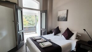 A hotel room with a bed, a nightstand, a lamp, a painting, and a balcony with a view of trees. - The Grand Hotel Newcastle (Newcastle)