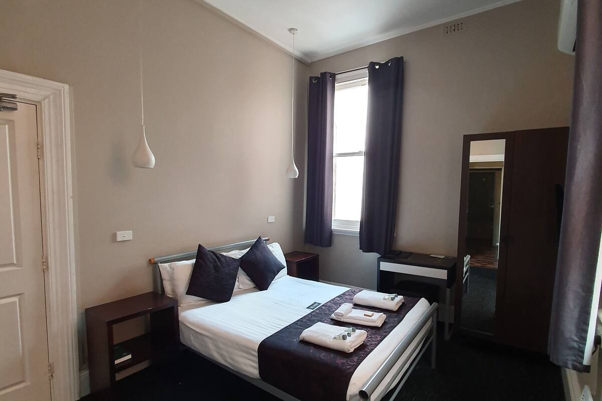 Habitación de hotel con cama, mesitas de noche, ventilador de techo, ventana con cortinas, espejo y puerta.
