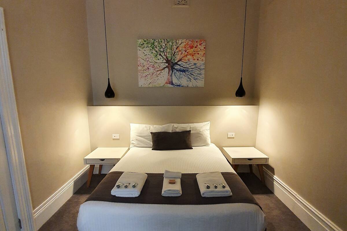 Una cama bien hecha con edredón blanco y cabecera marrón, flanqueada por dos mesitas de noche con lámparas, en una habitación con paredes color beige y una lámpara en el techo.