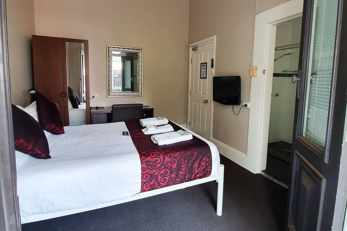 Habitación de hotel con cama, escritorio, espejo, televisor y zona de ducha.
