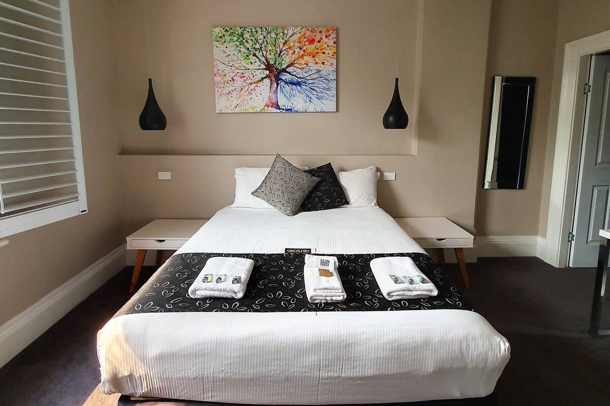 Una cama bien hecha con colcha de diseño blanco y negro, dos mesitas de noche, un cuadro de un árbol y una ventana con persianas.