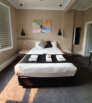 A neatly made bed with a black and white patterned bedspread, two bedside tables, a painting of a tree, and a window with blinds. - The Grand Hotel Newcastle (Newcastle)