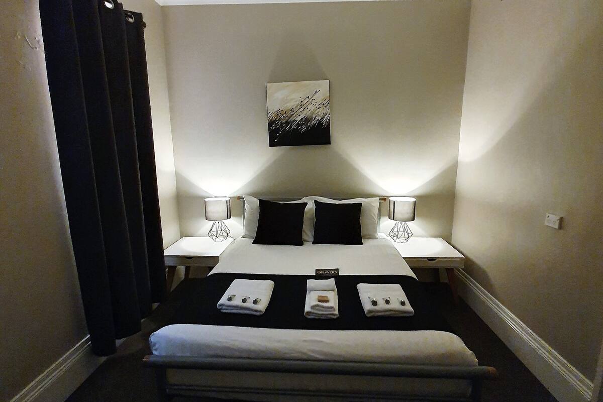 Habitación de hotel con cama, dos mesitas de noche, una obra de arte en la pared y una ventana con cortinas.