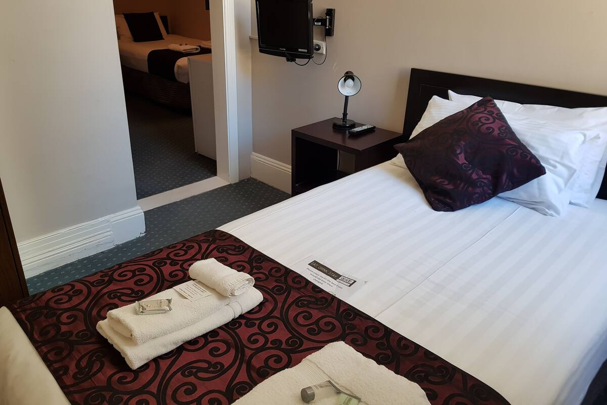 Una habitación de hotel con una cama bien hecha, una mesita de noche con lámpara, un televisor montado en la pared y una puerta que da a otra habitación.