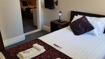 A hotel room with a neatly made bed, a bedside table with a lamp, a TV mounted on the wall, and a door leading to another room.