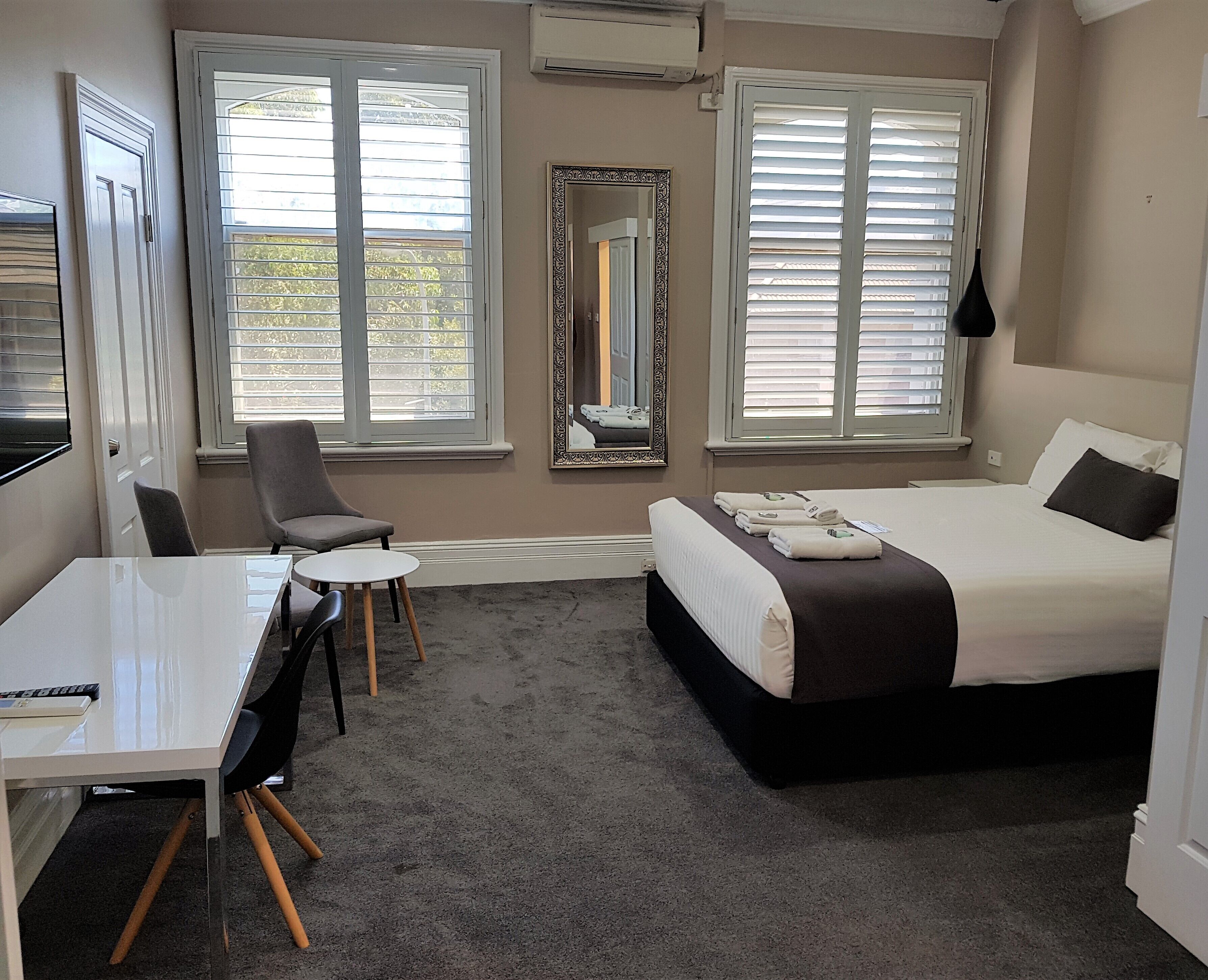 A hotel room with a bed, a desk, a chair, and two windows with blinds.