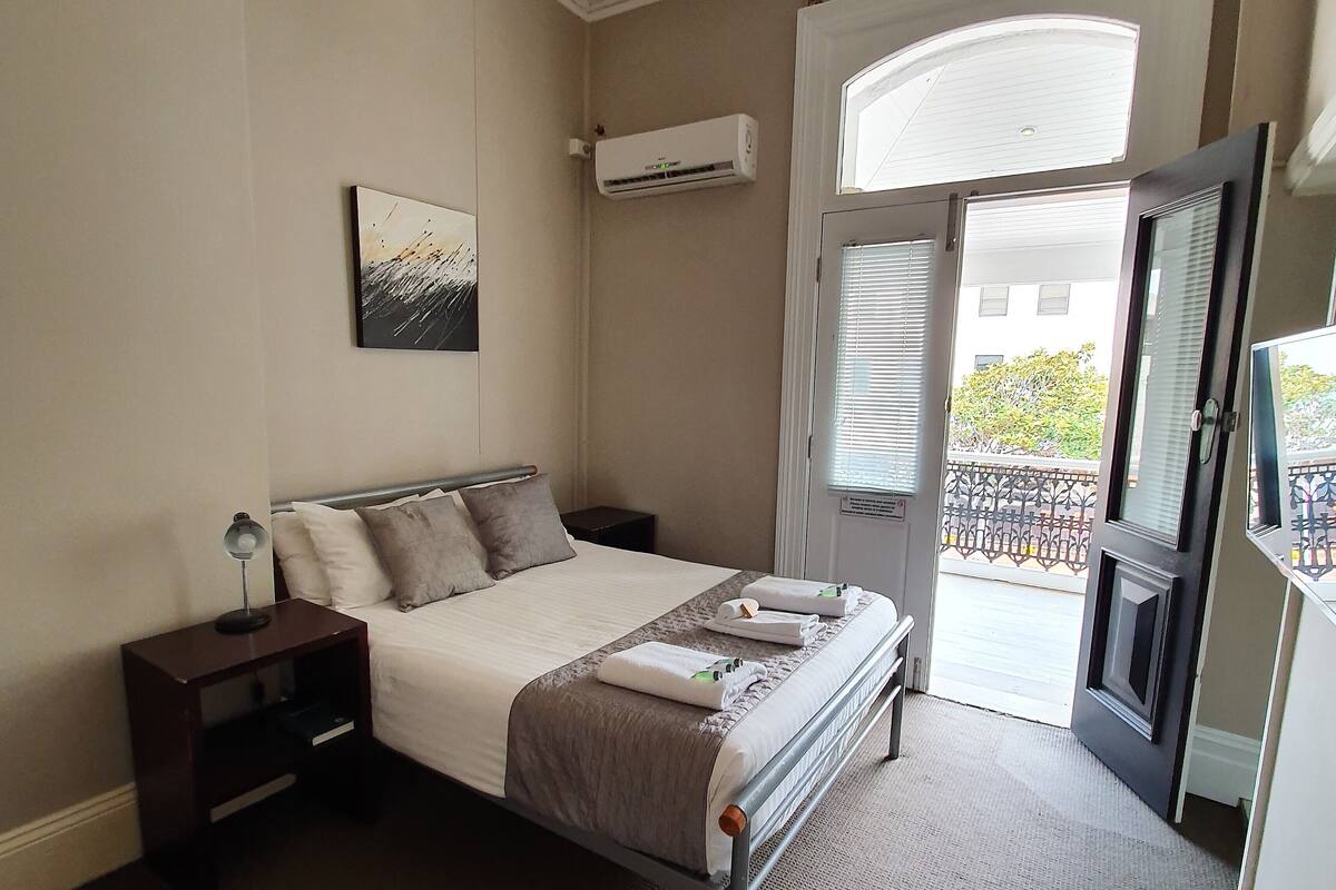 Un dormitorio con cama, mesitas de noche, aire acondicionado de pared, ventana con persianas y vista al balcón exterior.