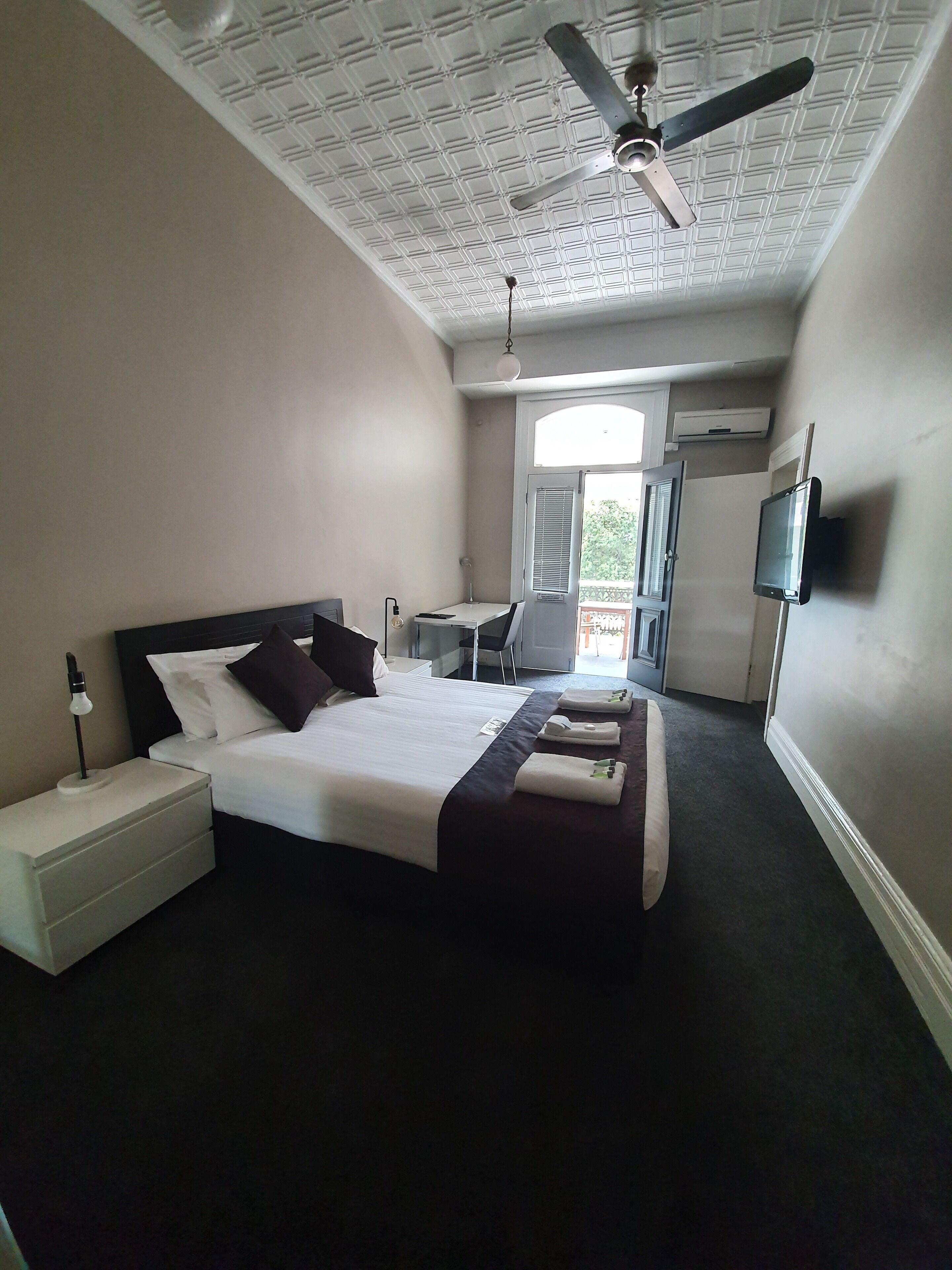 Een hotelkamer met een bed, nachtkastje, lamp, plafondventilator en een aan de muur bevestigde televisie.