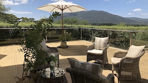 Una terraza de madera con mobiliario de mimbre y un amplio paraguas, con vistas a un paisaje montañoso.