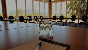 Una mesa de conferencias con un vaso, un bolígrafo y un bloc de notas en primer plano, y una vista de sillas y un paisaje a través de amplias ventanas al fondo.