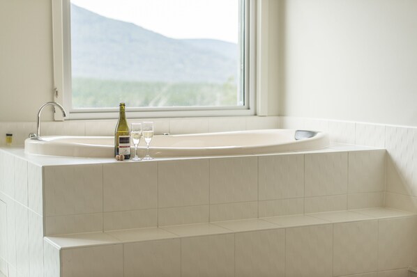 A bathroom with a large bathtub, a window with a view of mountains, and a bottle of wine and glasses on the sink.