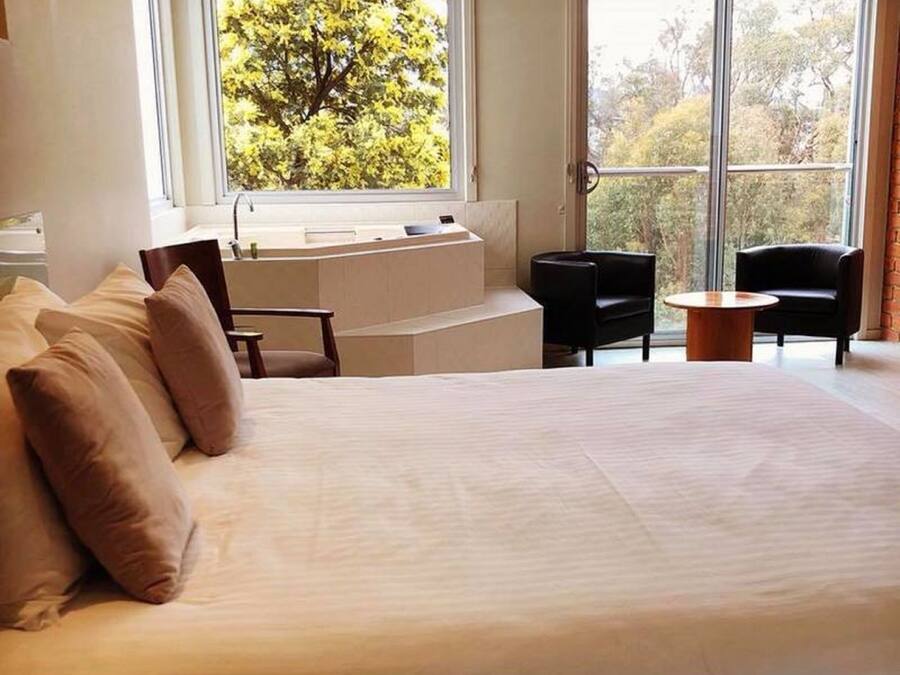 Una cama grande con vistas a árboles a través de una ventana. Hay una pequeña mesa y dos sillas en la habitación.